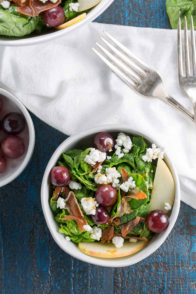 Top down view of a bowl of spinach salad with red pickled grapes, crumbled chèvre, crispy prosciutto bits, and sliced apple, next to a bowl of red grapes and a white cloth topped with two silver forks, on a blue wooden background, with a larger bowl of salad visible towards the top left edge of the frame.