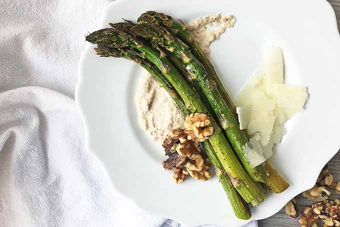 Horizontal image of a plate with asparagus, sauce, nuts, and cheese on a white towel.