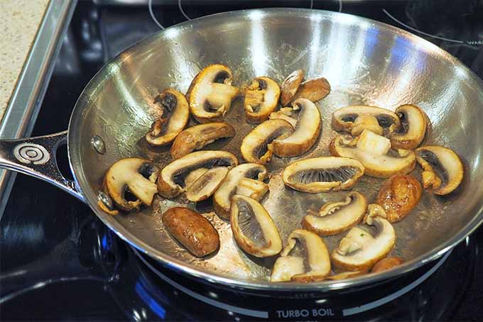 Top view of sliced mushrooms being browned in a large stainless steel frying pan.