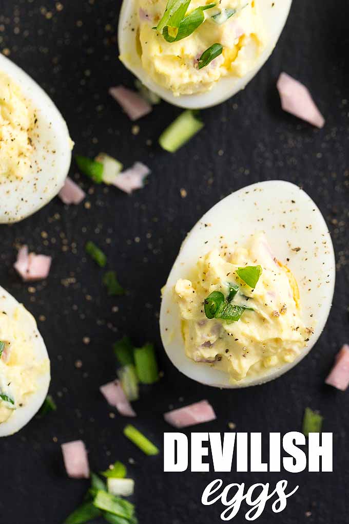 Top-down view of several halved hard-boiled eggs filled with a pale yellow yolk filling and topped with chopped chives, on a black background with scattered bits of ham and herbs, printed with the words "Devilish eggs" at the bottom right corner.