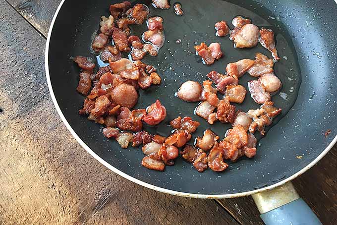 Horizontal image of a skillet on a wooden surface with cooked bacon pieces.