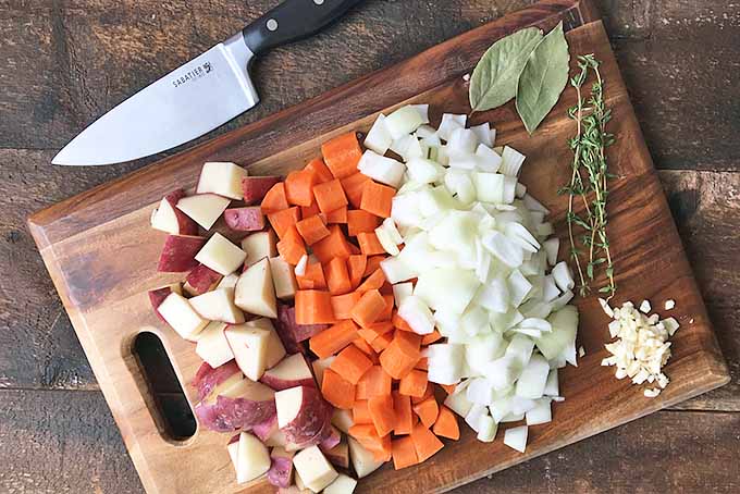 Horizontal top-down image of chopped vegetables and herbs on a wooden cutting board with a knife on the side. 
