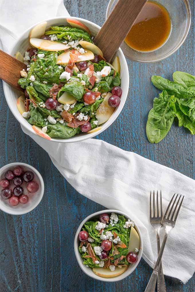 Top down view of a large white ceramic bowl of spinach salad with scattered leaves of spinach, a white ramekin of red pickled grapes, a smaller bowl of salad, two forks, and a white cloth on a blue wooden background.