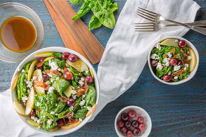 Top down view of a large white bowl of spinach salad with apples, prosciutto, goat cheese crumbles and red grapes, with a smaller bowl of salad to the right, a small white ramekin of pickled grapes, the handles of brown wooden serving utensils, and a few leaves of spinach, with a white cloth and two silver forks on a blue wooden background.