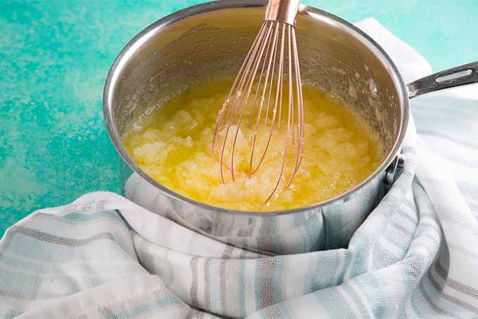 A stainless steel mixing bowl filled with a yellow mixture of melted butter and sugar, being stirred with a copper colored whisk, on a blue background with the base of the bowl wrapped in a white and gray striped dish towel.