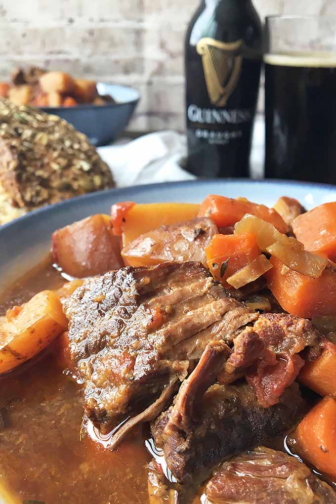 Vertical closeup image of beef stew in a bowl, with bread, beer, and another bowl of stew in the background on a wooden surface with a white towel. 