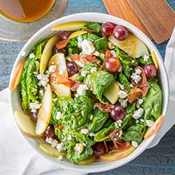 Square top down image of spinach salad in a white bowl with crumbled goat cheese, sliced apple, pickled grapes, crispy prosciutto, and balsamic vinegar dressing, on a blue wooden background with a white cloth napkin, wooden serving utensils, and a glass bowl of dressing.