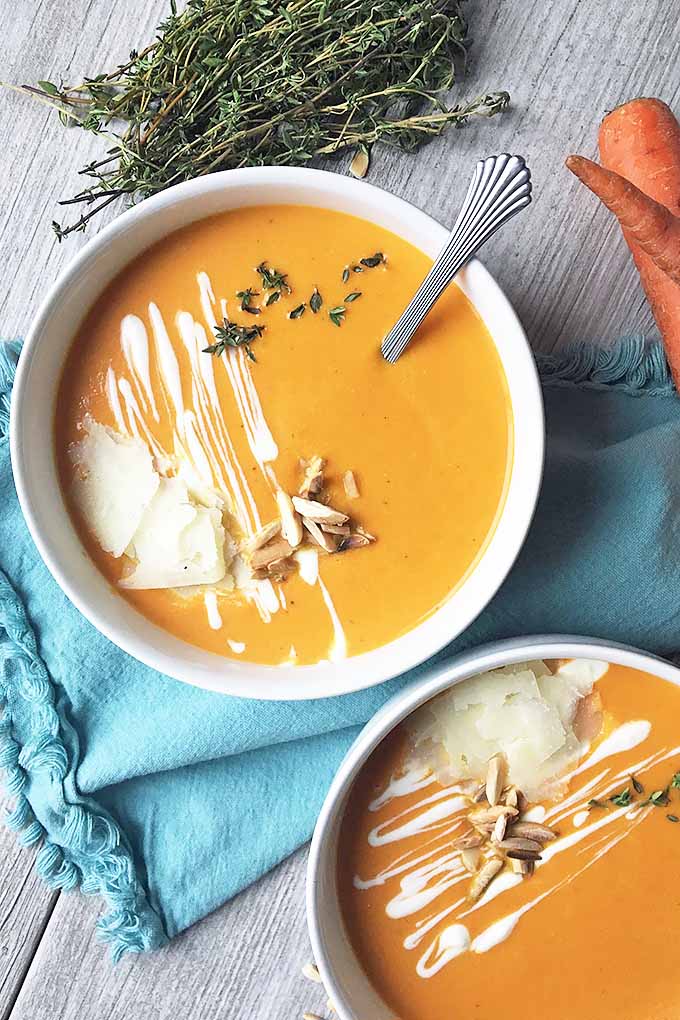 Vertical image of bowls of carrot soup with assorted garnishes and a metal spoon, with thyme, carrots, and a blue towel on a gray wooden surface.