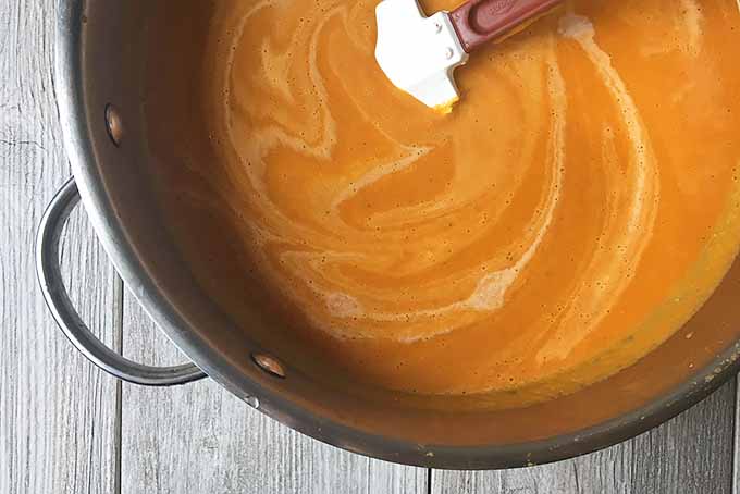 Horizontal image of a spatula stirring cream into an orange mixture in a metal bowl on a gray wooden surface.