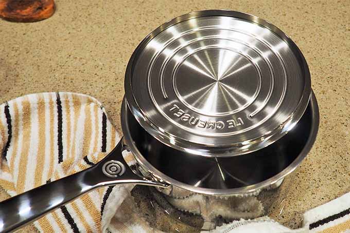 Bottom of a stainless steel pot lid resting in a saucepan, with concentric circles on the bottom of the lid, on a beige counter with a beige, black and white striped kitchen towel.