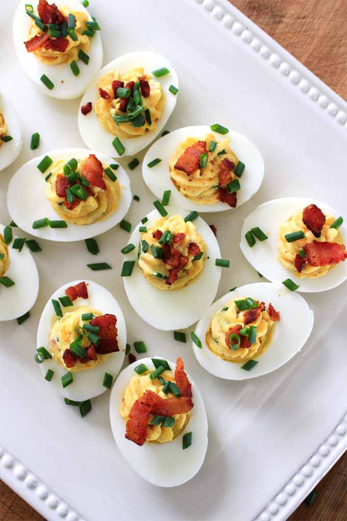 Vertical top-down view of 12 hard boiled egg halves arranged in rows, filled with a deviled yolk mixture, and topped with a garnish of bacon crumbled and chopped chives, on a rectangular white plate.