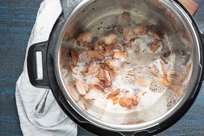 Top-down view of bacon cooking in the base of a slow cooker pot on the sear setting, with sizzling fat, and a white dish towel next to and wrapped around the base of the black and silver slow cooker.