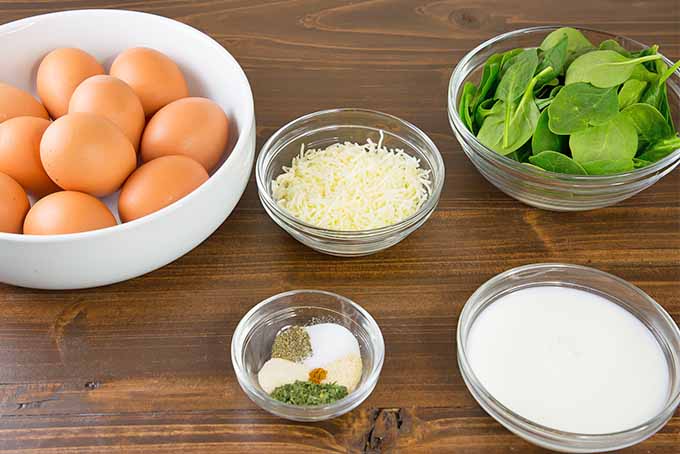 A white ceramic bowl of brown eggs, a glass dish of shredded cheese, a small glass bowl of salt and dry spices, a bowl of spinach, and a bowl of milk are arranged on a brown wooden surface.