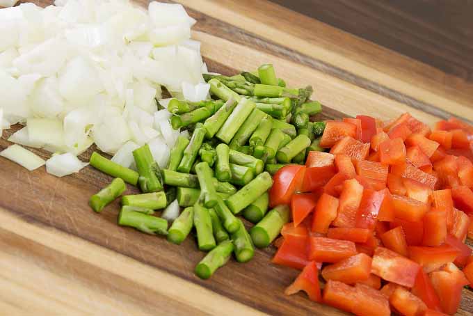 Three equally-sized piles of chopped white onion, asparagus, and red bell pepper on a striped wooden cutting board.