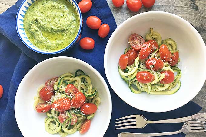 Horizontal image of two bowls with fresh zoodles and tomatoes with forks, a bowl of pesto, and whole tomatoes on a blue towel.