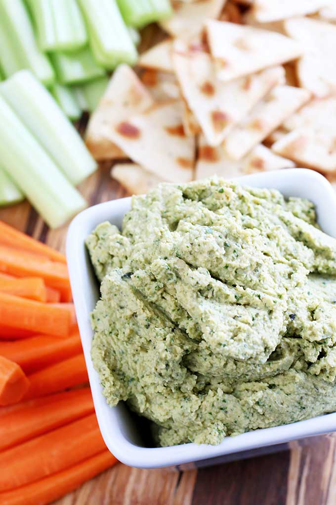 Vertical image of a white bowl with a kale hummus dip surrounded by pita and fresh vegetables.