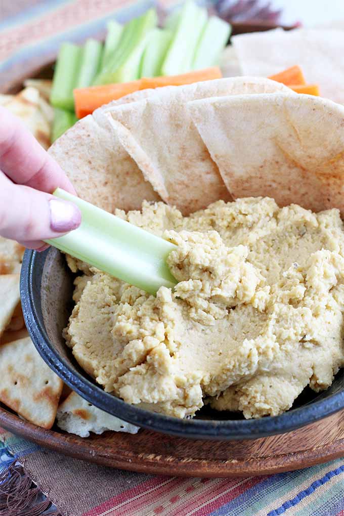 A woman's hand with shiny pink fingernails dips a stick of celery into a small bowl of homemade hummus, with pita bread and chips, celery sticks, and carrot sticks on a wooden plate, on a striped fabric background.