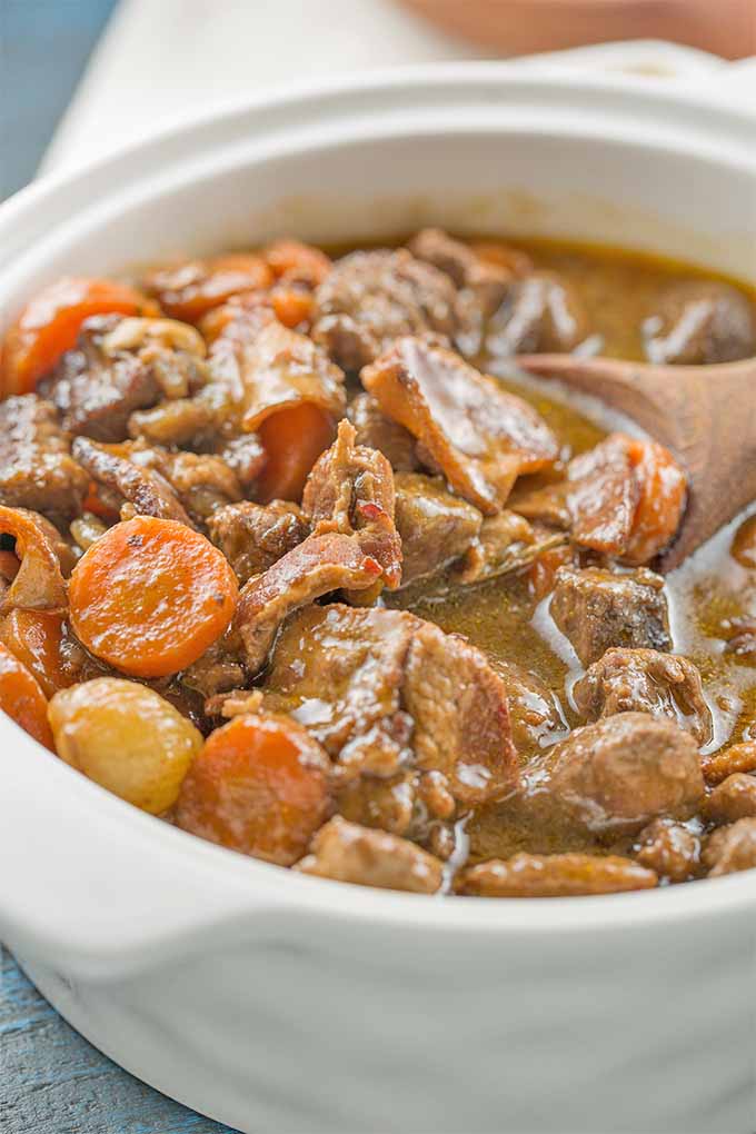 Vertical image of a white ceramic serving dish full of slow cooked beef bourguignon, with chunks of beef, sliced carrots, pearl onions, and criminology mushrooms, with a wooden spoon stuck into the dish.