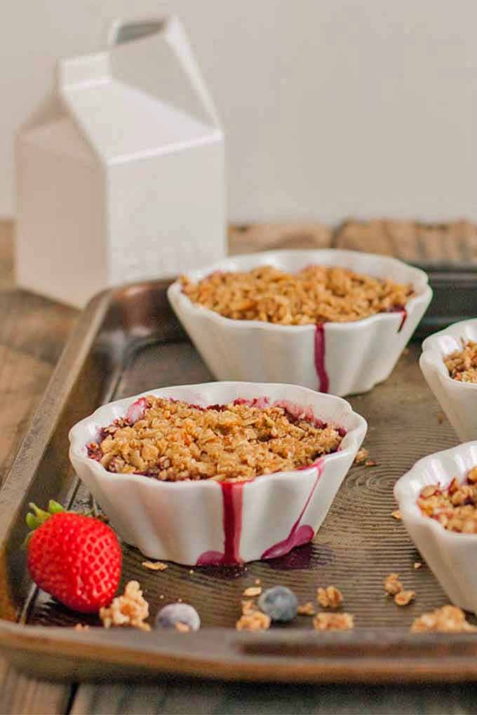 Vertical image of four fluted custard cups of berry crumble with red juice dripping down the sides, with scattered crumble and berries on a baking sheet, on top of a brown table against a beige wall, with a ceramic milk carton creamer in the background.
