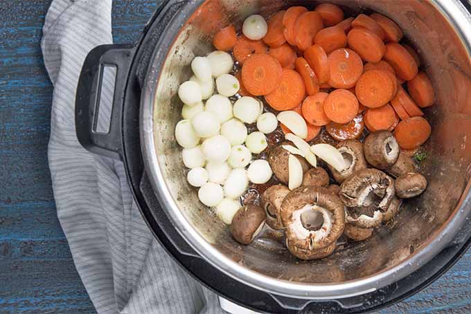 Top-down view of raw sliced carrots, pearl onions, and mushrooms in a slow cooker, with a towel wrapped around the slow cooker base.