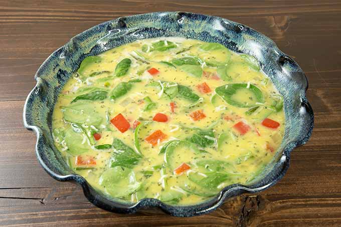 A blue-green ceramic pie dish is filled with raw beaten egg mixed with chopped sautéed bell pepper, onion, and asparagus, and fresh spinach, on a brown wooden table.