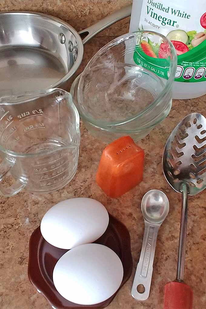 Vertical image of a stainless steel frying pan, two small glass custard dishes, a plastic jug of white distilled vinegar, a metal slotted spoon, a metal teaspoon, an orange salt shaker, two white eggs on a brown coaster, and a glass pitcher-style measuring cup, on a tan speckled countertop.