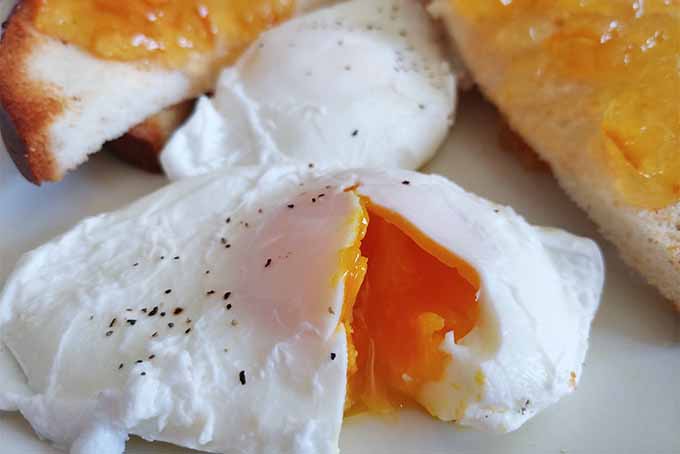 Closeup of a poached egg sprinkled with black pepper and broken open to show a vibrant orange soft-cooked center, on a white plate with another poached egg and white toast topped with orange marmalade.