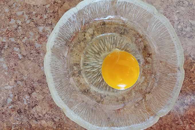 Top-down view of a raw egg cracked into a small glass bowl, on a tan speckled granite background.