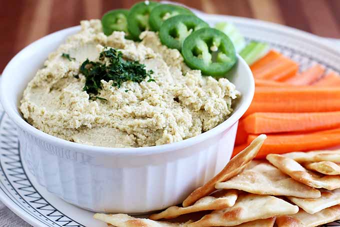 Horizontal image of a white dish filled with freshly made hummus and sticks of carrots and pita chips. 