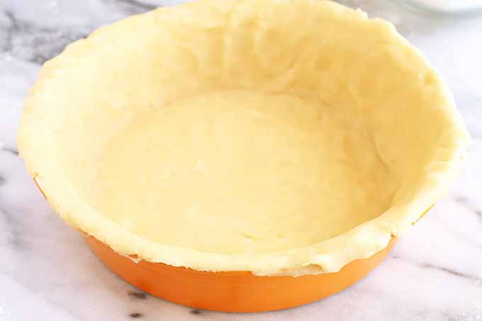 An orange ceramic pie plate is lined with a buttery pie crust, on a white and gray piece of marble.