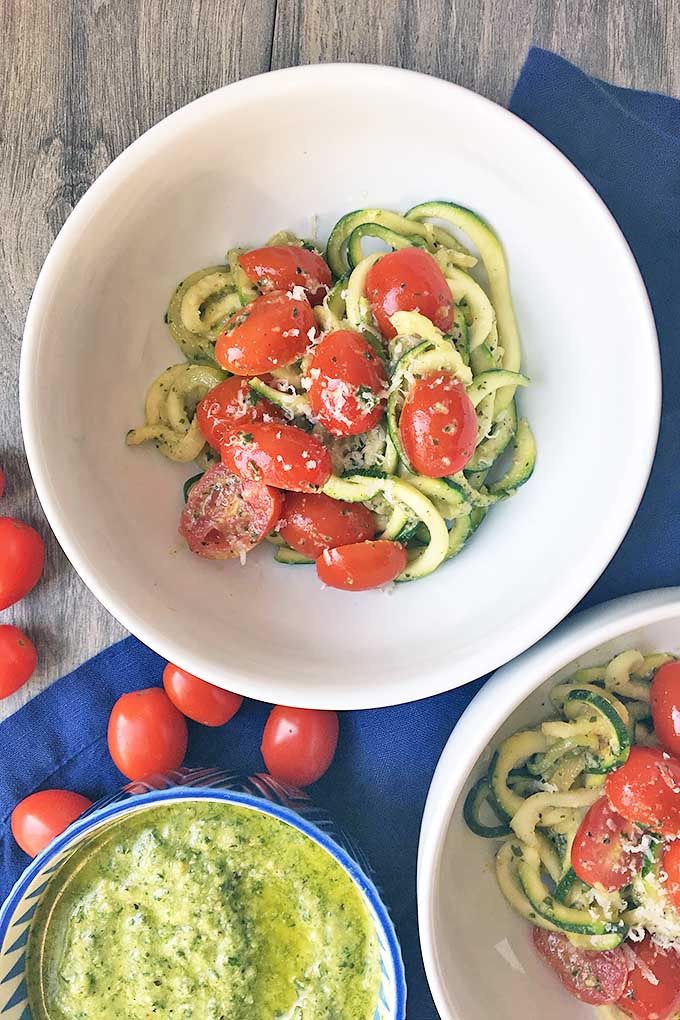 Vertical image of white bowls with zucchini noodles and tomatoes on a blue towel, with whole tomatoes and a bowl of pesto next to them. 