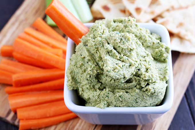 Horizontal image of a carrot stick dipped into a white bowl of fresh green hummus.