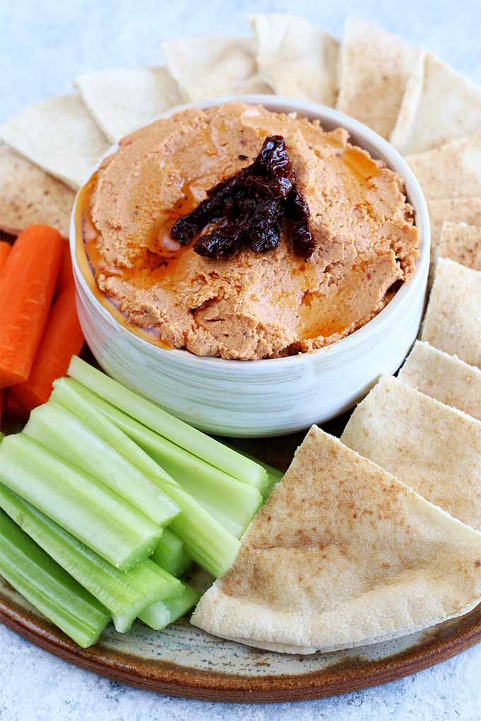 Vertical image of a beige platter topped with carrot sticks, celery sticks, triangles of hummus, and a small bowl of light orange hummus topped with chopped sun-dried tomatoes, on a pale blue background.