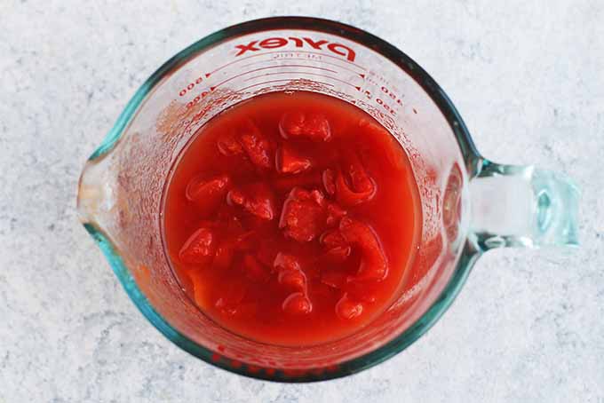 Top-down view of a glass Pyrex pitcher-style liquid measuring cup filled with diced tomatoes and their juices, on a white background.