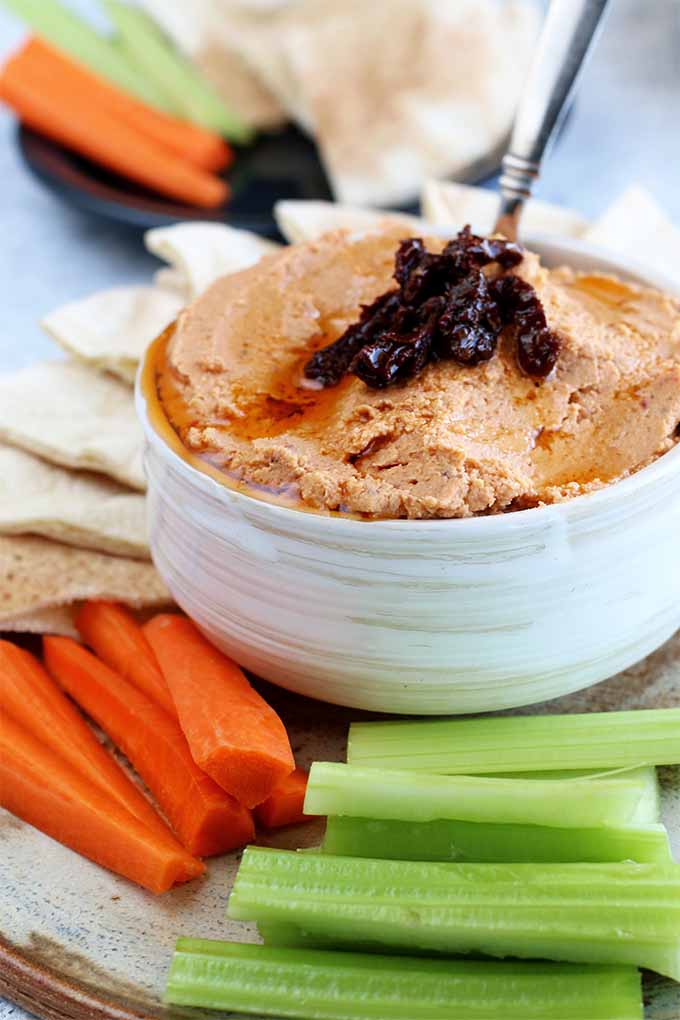 Vertical image of a white ceramic bowl filled with sun-dried tomato hummus, on a round beige platter of celery sticks, carrot sticks, and sliced pita bread arranged around the dip bowl, with a navy blue plate of vegetables, hummus,. and pita bread in shallow focus in the background.
