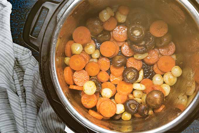 Top-down image of cooked sliced carrots, pearl onions, and button mushrooms in a slow cooker, with steam slightly obscuring the view into the pot.