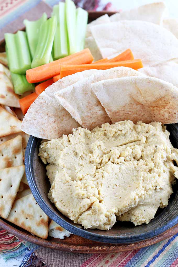 Vertical image of a small black bowlful of homemade hummus with fanned out slices of pita, with more pita bread and chips, celery, and carrot sticks, arranged on a serving platter, on top of a patterned pink and purple cloth place mat.