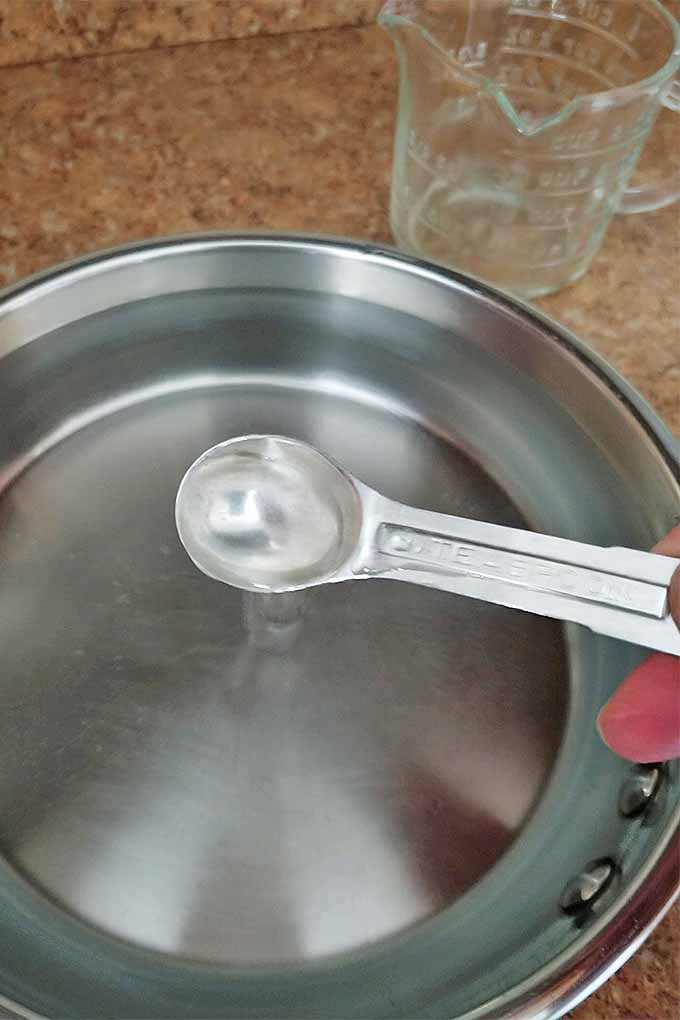A hand pours a metal teaspoon of white vinegar into a frying pan of water, on a tan granite countertop beside a glass measuring pitcher.