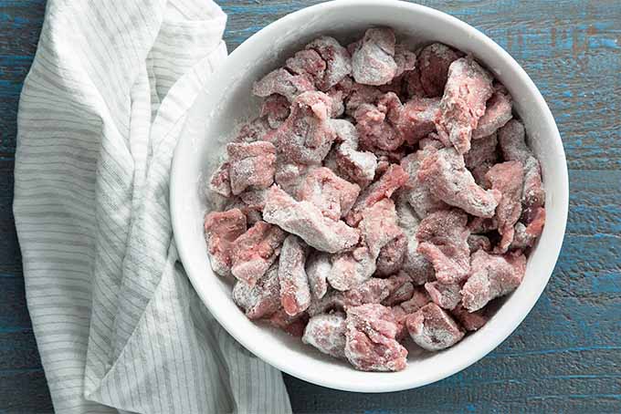 Top-down view of piece of beef stew meat dredged in seasoned flour in a white bowl, next to a crumpled white kitchen towel on a blue-gray wooden background.