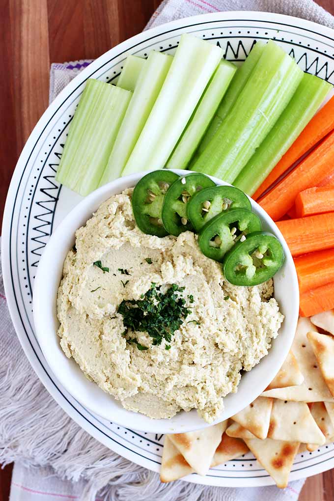 Vertical top-down image of a bowl of hummus dip with cilantro and jalapeno, with celery, carrots, and pita chips. 