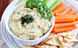 Horizontal image of hummus garnished with jalapeno and cilantro, on a platter with celery, carrots, and pita triangles.