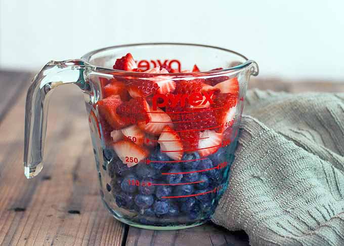 A glass Pyrex measuring cup filled with blueberries and sliced strawberries, on a brown rustic wooden table in front of tan wall, beside a moss green dish towel.