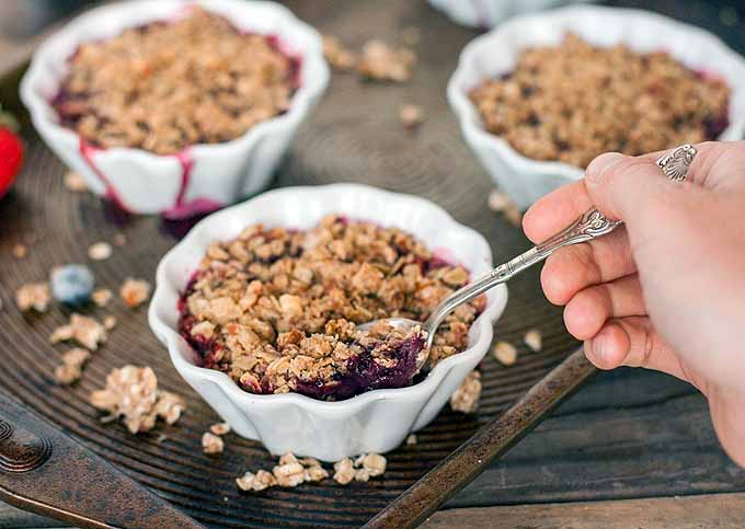 A hand digs a spoon into a custard cup of berry crumble, beside two other cups of crumble dessert on a baking sheet with scattered oat and almond crumble clumps and berries, on a brown wooden surface.