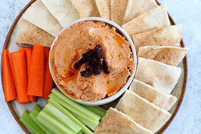 Top-down view of a dip bowl filled with light orange sun-dried tomato hummus, on a platter surrounded with fanned out pita bread sliced into triangles, orange carrot sticks, and pale green celery sticks, on a mottled pale blue background.