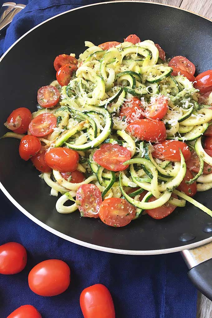Vertical image of a large pan with a mixture of pesto, zoodles, and chopped tomatoes.