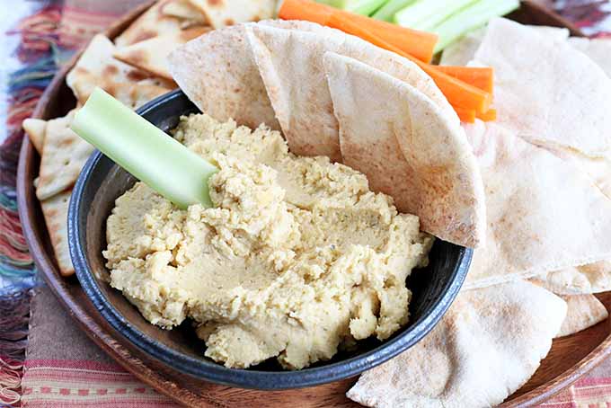 An appetizer platter of crudite, pita bread and chips, and a small navy blue bowl of beige garlic hummus, with a celery stick dipped into the spread.
