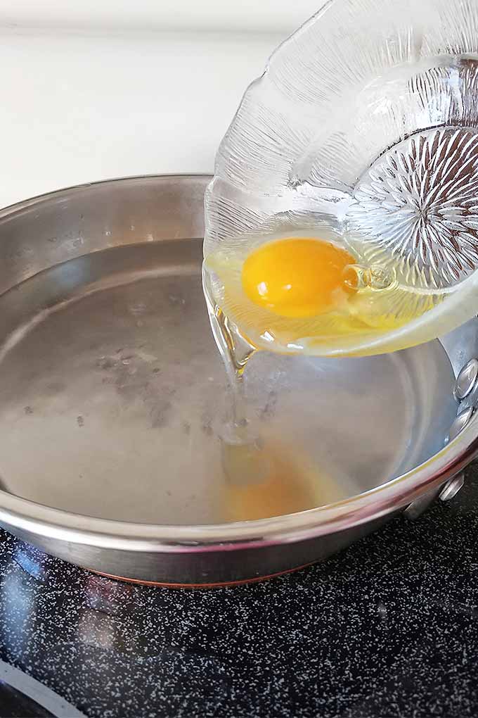Vertical image of a small glass dish with a raw egg in it being poured into a stainless frying pan filled with an inch of water, on a black flat-topped electric stove with a white wall in the background.