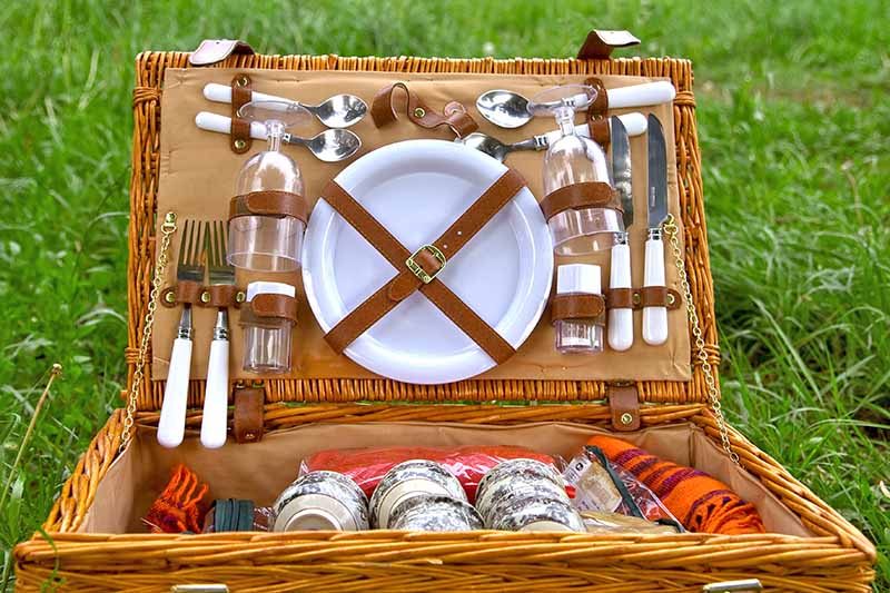 Horizontal closely cropped image of a fully stocked wicker picnic basked with glasses, silverware, dishes, and more, on a green lawn.