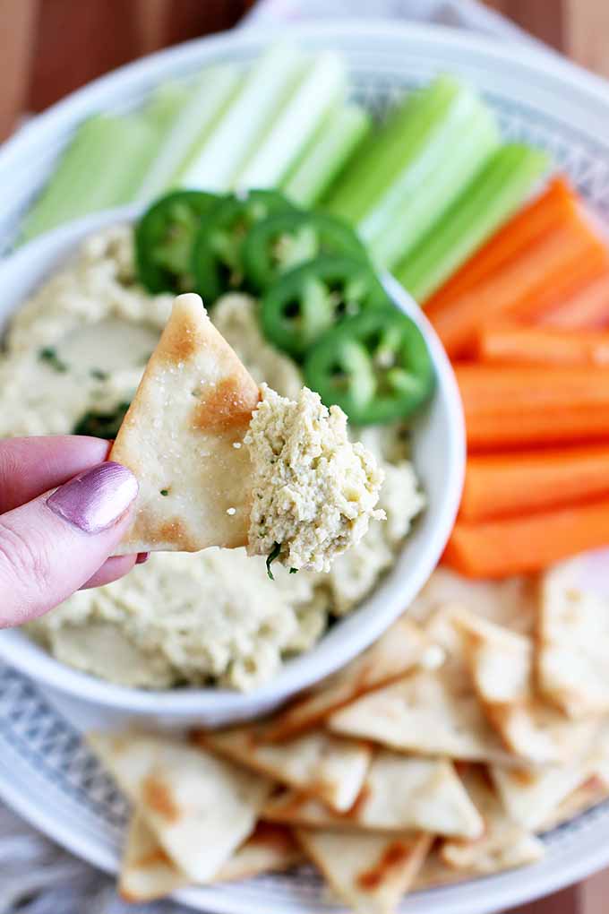 Horizontal image of a hand holding a pita chip dipped in hummus. 