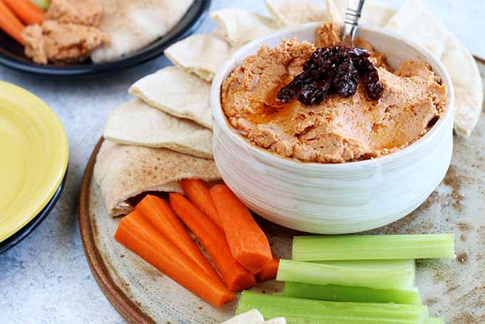 A small white bowl of sun-dried tomato hummus topped with sun-dried tomatoes, on a platter surrounded by triangles of pita bread, carrot sticks, and celery sticks, on a gray table beside a blue and yellow plate, and another dark blue plate of hummus and vegetables.